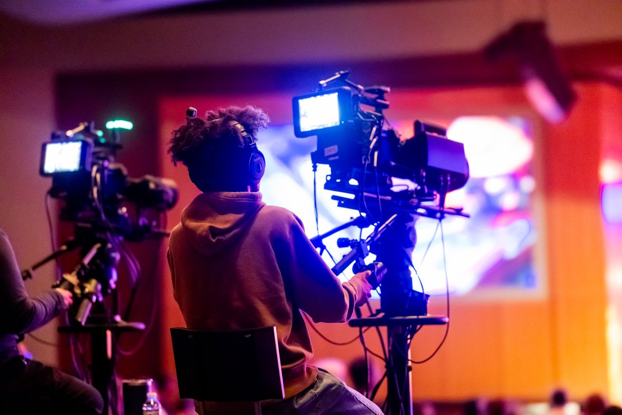 Contact Back view of a videographer operating a camera at a live event, focusing on a vibrant indoor setup.
